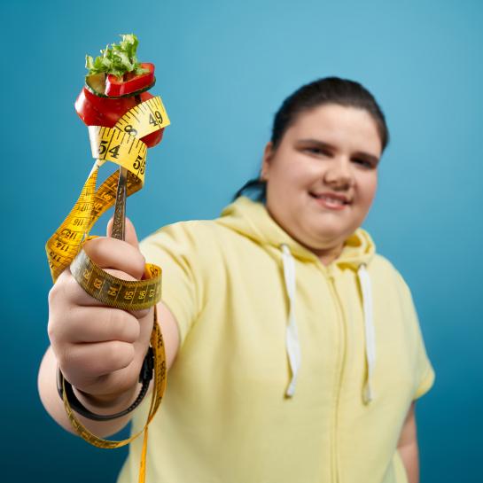 Plump smiling teen girl holding fork with vegetables and measuring tape wrapped around it