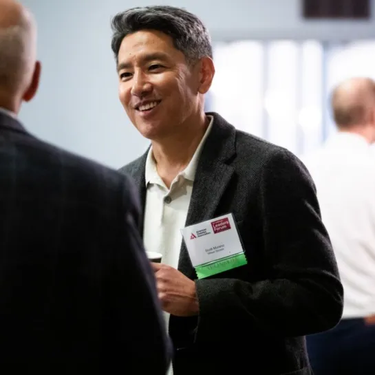 Man in jacket and name tag smiles, talking to colleague at a conference.