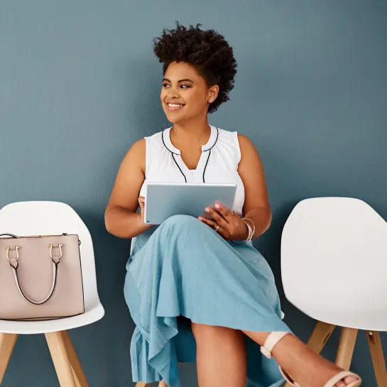 Smiling Black woman in white top and blue skirt sits holding a tablet, looking left.