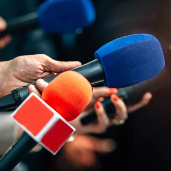 Photograph: Hands holding reporter microphones with blue and orange foam covers.