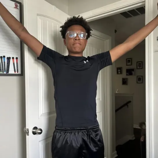 Young man in black athletic wear and glasses stands with arms outstretched.
