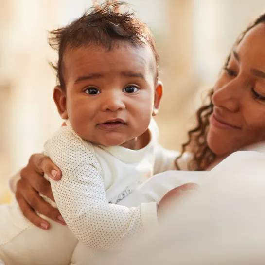 Happy mother cradles baby in white outfit; baby looks at camera with curious eyes.