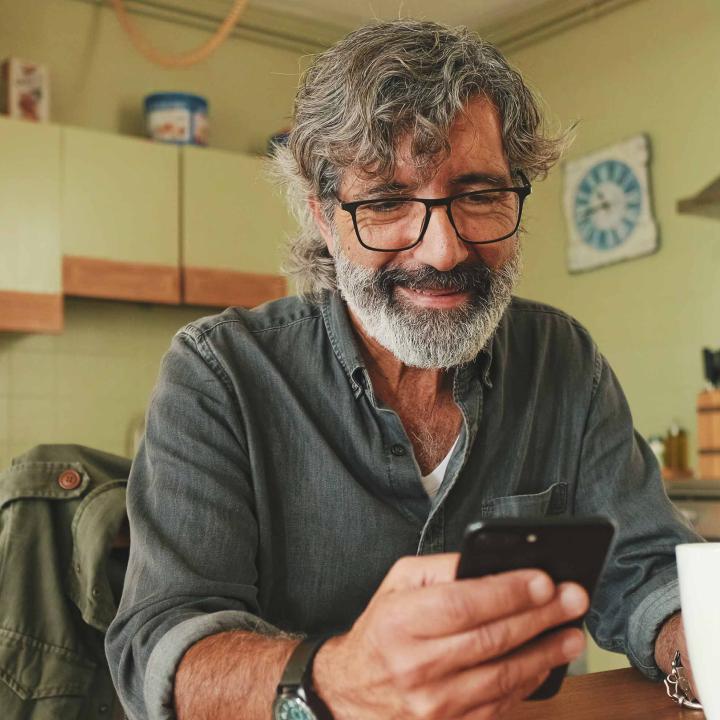 Senior latino man beard and glasses drinking coffee and looking at phone