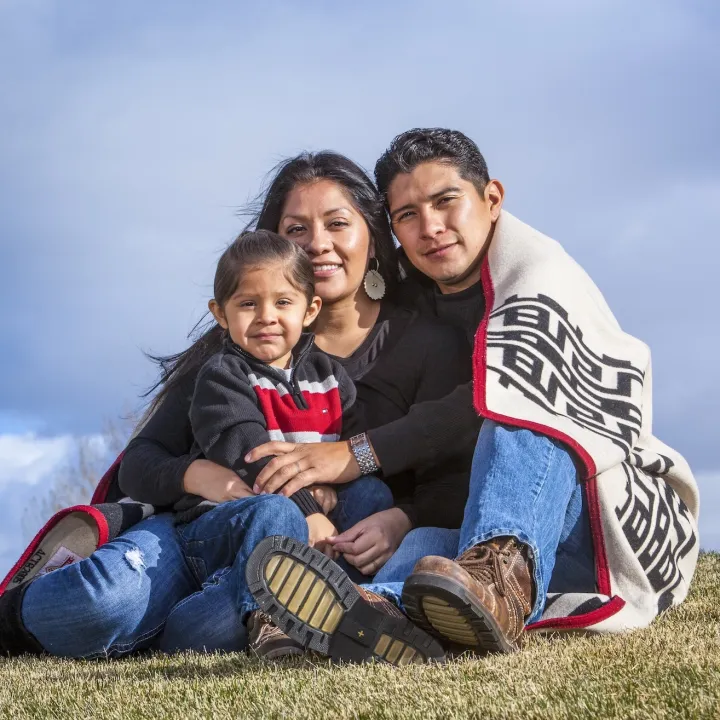 Smiling Indigenous family of three sits on grass; dad wrapped in a patterned blanket.
