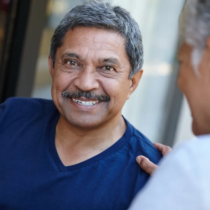 Smiling man with gray hair and mustache, wearing blue, with a hand on his shoulder.