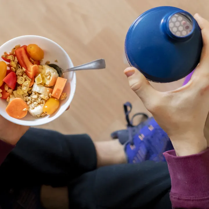 Hands hold a bowl of colorful fruit and grain with a blue water bottle.