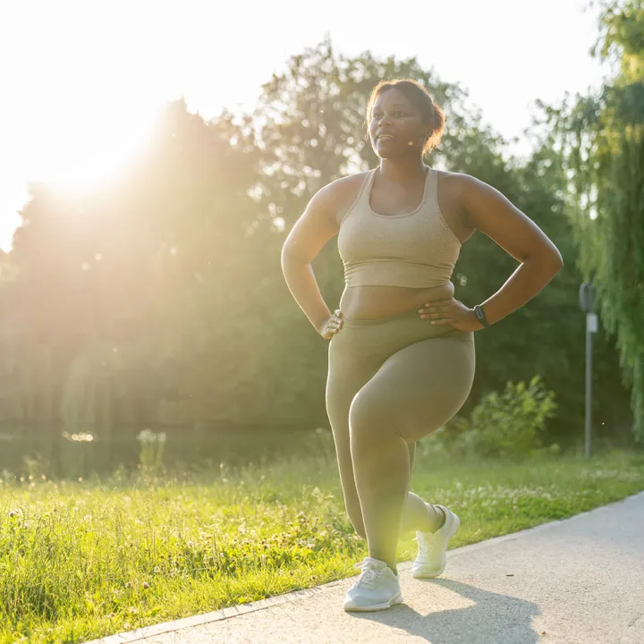 Black woman doing a lunge outdoors in a sunny park.
