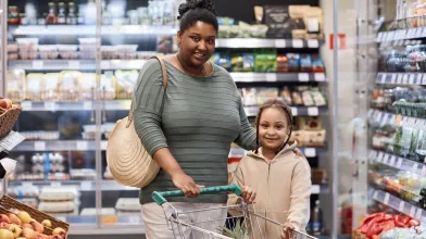 A Black mom and daughter pushing a shopping cart in a supermarket aisle filled with refrigerated food.