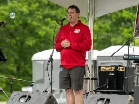 Man in red sweatshirt and shorts speaks into a microphone on an outdoor stage.