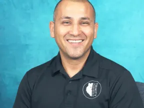 Smiling man in a black polo shirt against a textured blue background.