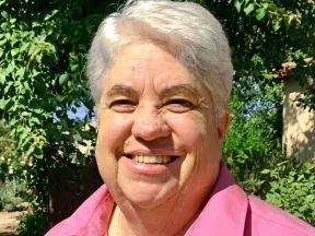 Smiling person with white hair in a pink shirt, outdoors with green plants.