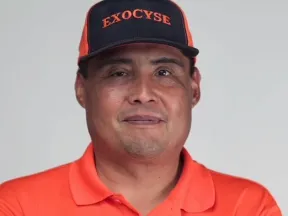 Smiling man in orange polo and blue "EXOCYSE" cap with crossed arms.