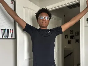 Young man in black athletic wear and glasses stands with arms outstretched.