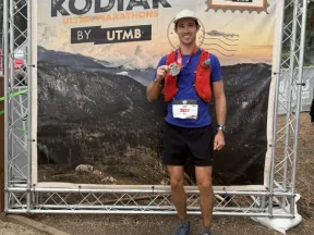 Smiling man holds race medal in front of "HOKA Kodiak Ultra-Marathon" banner.