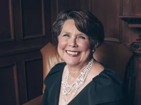 Smiling woman in black dress and pearls seated in leather armchair, wood-paneled room.