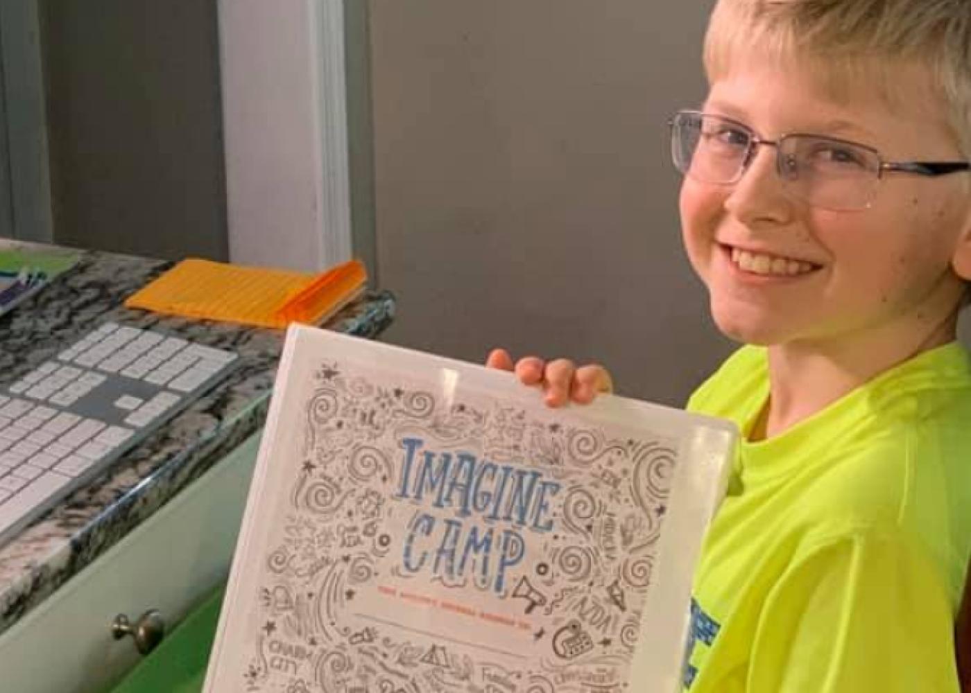 Blond boy with glasses in yellow shirt holding imagine camp piece of paper