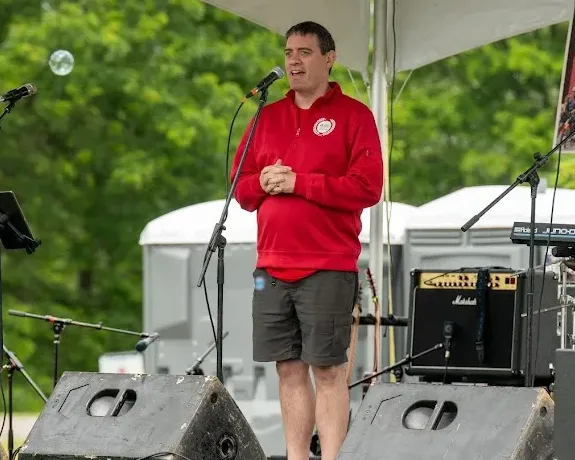Man in red sweatshirt and shorts speaks into a microphone on an outdoor stage.