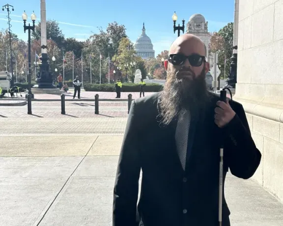 Blind man with beard, sunglasses, and white cane outside the U.S. Capitol.