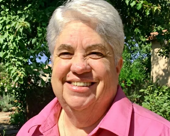 Smiling person with white hair in a pink shirt, outdoors with green plants.