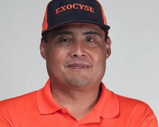 Smiling man in orange polo and blue "EXOCYSE" cap with crossed arms.