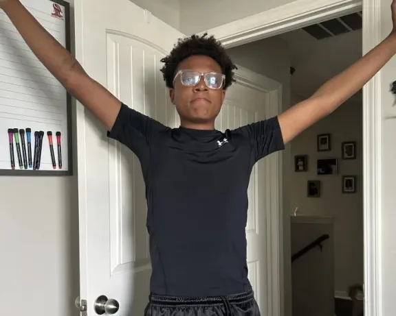 Young man in black athletic wear and glasses stands with arms outstretched.