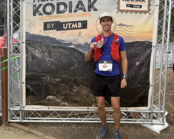 Smiling man holds race medal in front of "HOKA Kodiak Ultra-Marathon" banner.