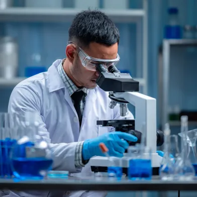 Scientist in lab coat and goggles examines a sample under a microscope in a laboratory.