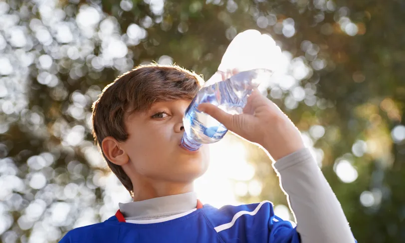 Due to having diabetes and high ketones, a young boy in blue shirt is drinking water from a bottle in sunlight.
