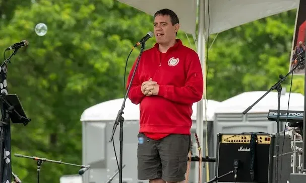 Man in red sweatshirt and shorts speaks into a microphone on an outdoor stage.