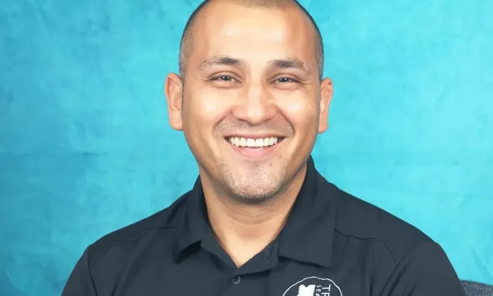 Smiling man in a black polo shirt against a textured blue background.