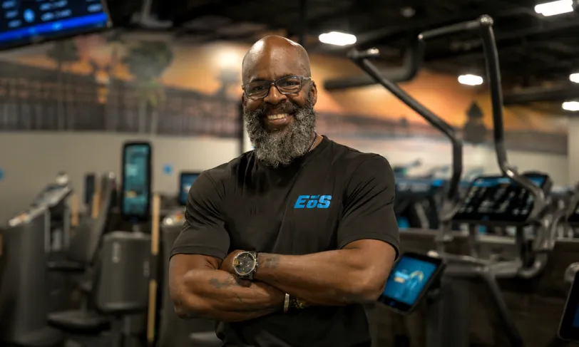 Confident man with beard smiles, arms crossed, in a modern gym.