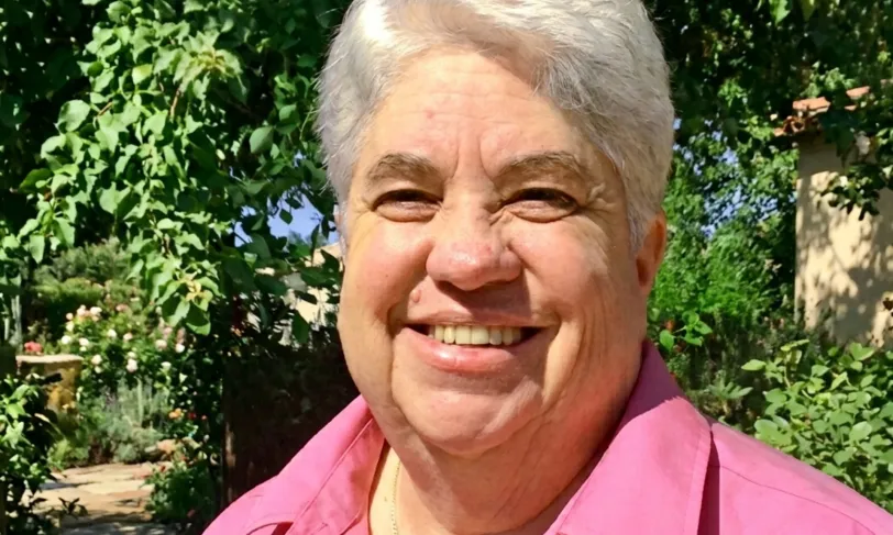 Smiling person with white hair in a pink shirt, outdoors with green plants.