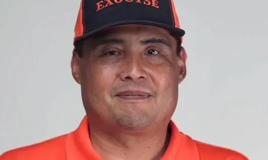 Smiling man in orange polo and blue "EXOCYSE" cap with crossed arms.