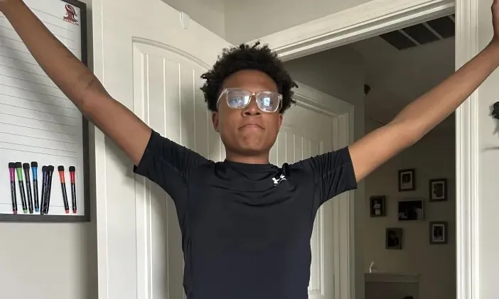 Young man in black athletic wear and glasses stands with arms outstretched.