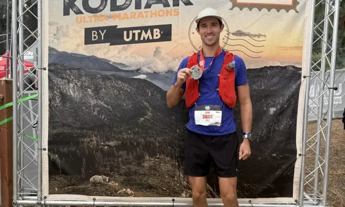 Smiling man holds race medal in front of "HOKA Kodiak Ultra-Marathon" banner.