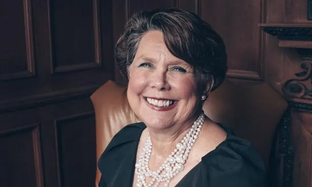 Smiling woman in black dress and pearls seated in leather armchair, wood-paneled room.