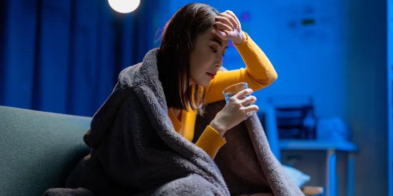 Photograph: Woman on couch, wrapped in blanket, holding head and water glass, looking distressed.