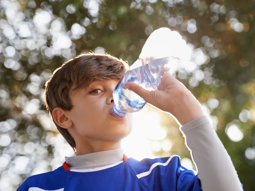 Due to having diabetes and high ketones, a young boy in blue shirt is drinking water from a bottle in sunlight.