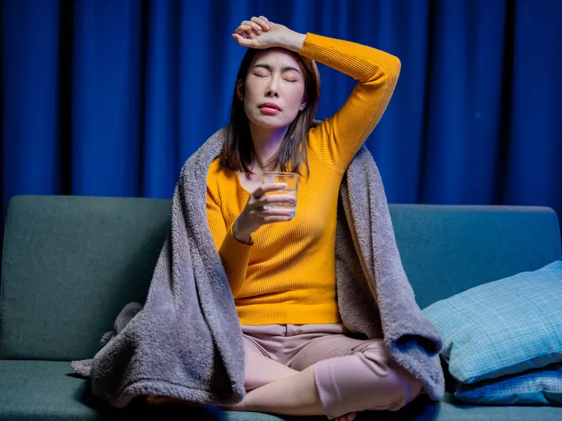 Unwell woman on couch, wrapped in blanket, hand to forehead, holding glass of water.