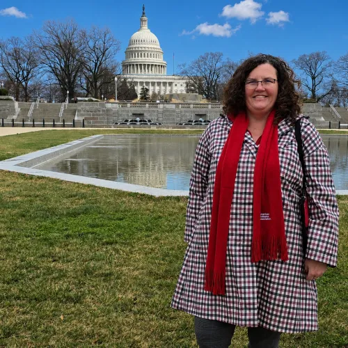 Smiling woman in plaid coat and red scarf stands before the U.S. Capitol.