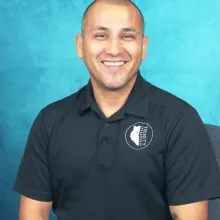 Smiling man in a black polo shirt against a textured blue background.