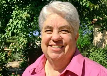 Smiling person with white hair in a pink shirt, outdoors with green plants.