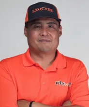 Smiling man in orange polo and blue "EXOCYSE" cap with crossed arms.