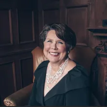 Smiling woman in black dress and pearls seated in leather armchair, wood-paneled room.