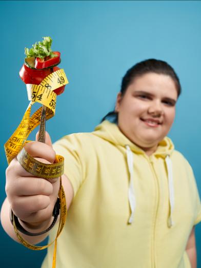 Plump smiling teen girl holding fork with vegetables and measuring tape wrapped around it