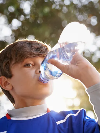 Due to having diabetes and high ketones, a young boy in blue shirt is drinking water from a bottle in sunlight.